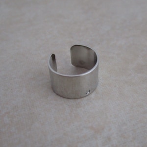 May include: A silver-colored cuff ring with a simple, modern design. The ring is open on one side and has a small hole on the outer surface. The ring is made of a shiny, metallic material.