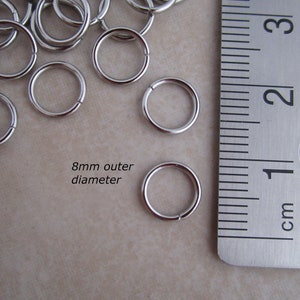 Puede incluir: Primer plano de anillos de salto plateados, que miden 8 mm de di&aacute;metro exterior, junto a una regla. La regla muestra medidas en cent&iacute;metros.