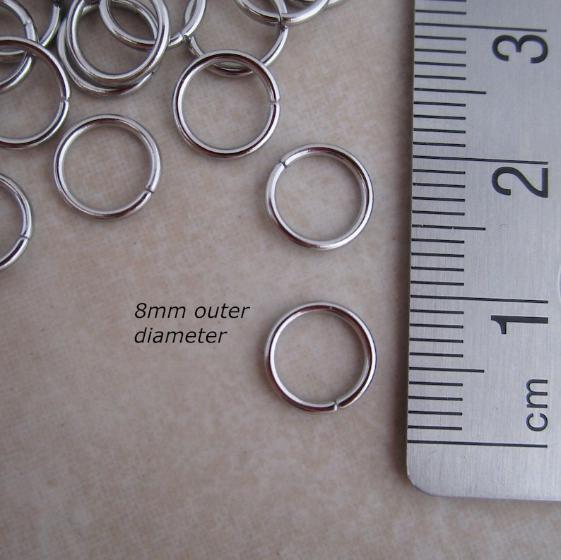 Puede incluir: Primer plano de anillos de salto plateados, que miden 8 mm de di&aacute;metro exterior, junto a una regla. La regla muestra medidas en cent&iacute;metros.