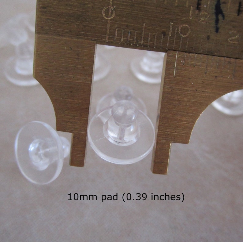 May include: Close-up of a clear plastic earring back with a 10mm pad, measured with a ruler. The text "10mm pad (0.39 inches)" is visible below the ruler.