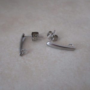 May include: Two silver-colored, curved bar earrings with a post and a small loop at the end. The earrings are made of a shiny metal and are displayed on a light-colored surface.