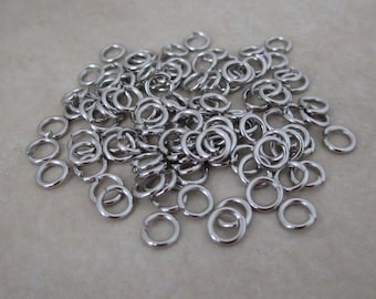 May include: A pile of small, shiny, silver-colored jump rings. These metal rings are open and ready to be used for jewelry making, crafts, or other creative projects. The rings are scattered on a neutral-toned surface.