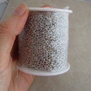 May include: A roll of silver chain link, wrapped in clear plastic, is held in a hand. The chain is wound around a white plastic spool.