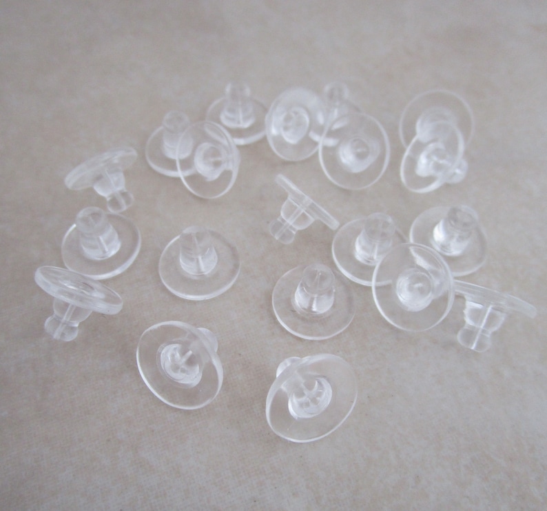 May include: A close-up of a group of clear plastic earring backs. The backs are small and round, with a small post in the center. They are designed to be used with stud earrings.