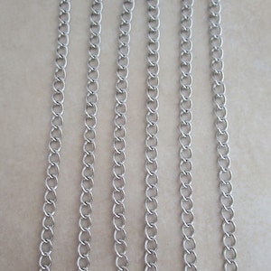 May include: Five strands of silver chain, each approximately 18 inches long, are laid out in parallel lines on a light beige surface.