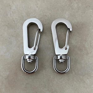 May include: Two silver-toned metal swivel lobster claw clasps with a polished finish. Each clasp features a spring-loaded gate mechanism and a rotating D-ring base, designed for attaching straps or accessories to bags, keychains, or other items.