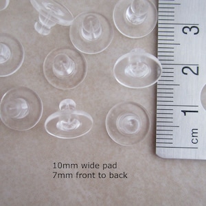 May include: A close-up of a group of clear plastic earring backs. The backs are 10mm wide and 7mm thick. They are arranged on a white surface next to a ruler.