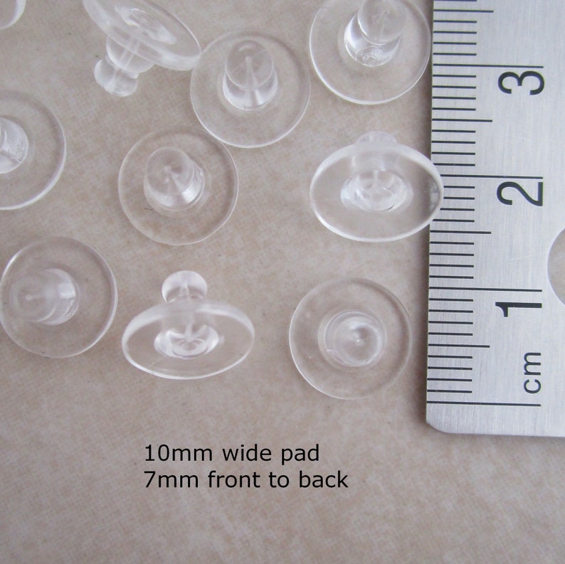 May include: A close-up of a group of clear plastic earring backs. The backs are 10mm wide and 7mm thick. They are arranged on a white surface next to a ruler.