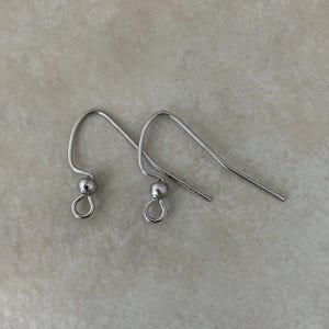 May include: A pair of silver-toned metal earring hooks featuring a curved wire design, a small decorative bead, and a circular loop at the bottom for attaching jewelry components. These findings are designed for creating dangling earrings.