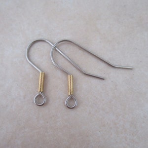 May include: Two silver-colored earring hooks with a gold-colored coil and a small loop at the bottom. The hooks are designed to be inserted into the ear and are suitable for creating handmade earrings. The hooks are displayed on a light beige surface.