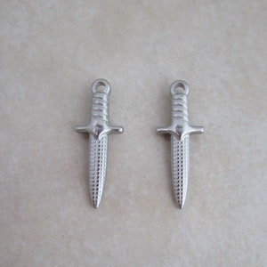 May include: Two silver dagger charms with a textured surface. The charms have a loop at the top for attaching to a necklace or bracelet.
