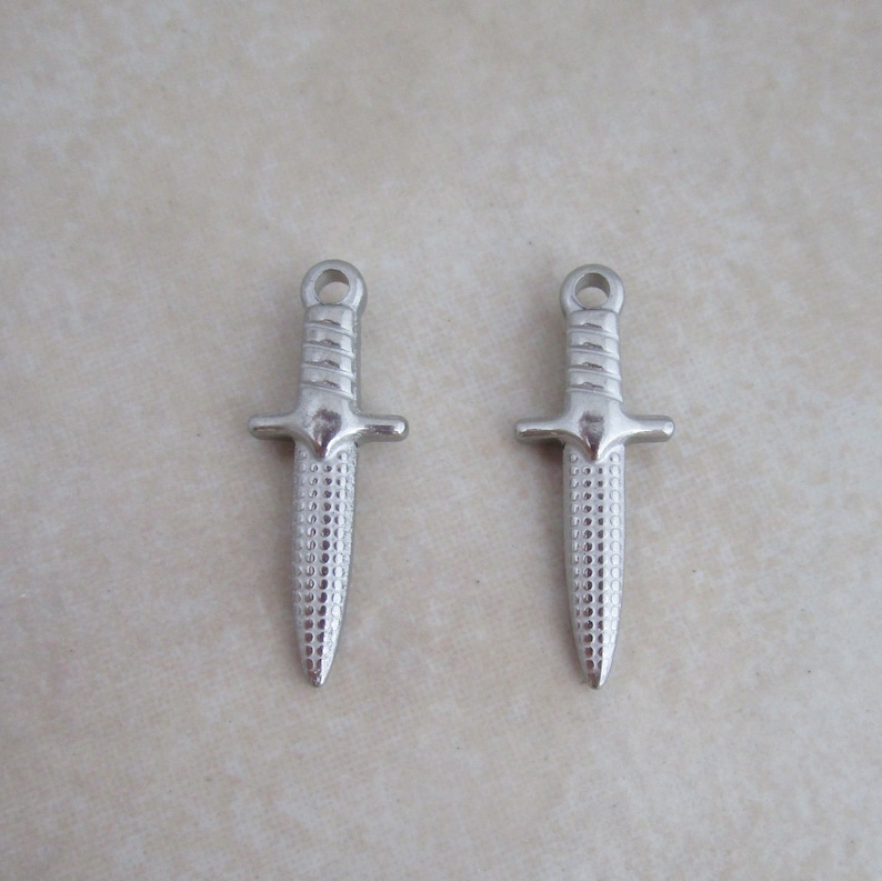 Op de afbeelding: Twee zilveren dolkbedels met een getextureerd oppervlak. De bedels hebben een lus aan de bovenkant om aan een ketting of armband te bevestigen.