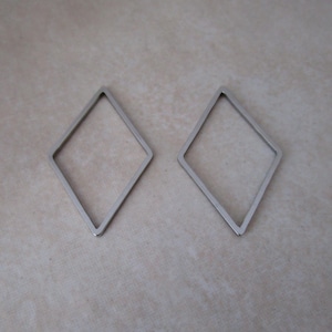 May include: Two silver-colored, open-frame diamond shapes. The geometric shapes are identical and lie flat against a neutral, textured surface. The diamonds are made of a thin, metallic material.