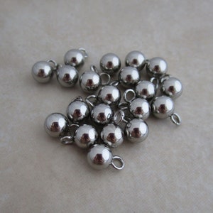 May include: A group of silver metal ball-shaped buttons with a loop on top. The buttons are arranged in a pile on a light gray surface.