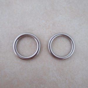 May include: Two silver metal rings, each with a smooth, circular shape. The rings are positioned side-by-side on a light beige surface.