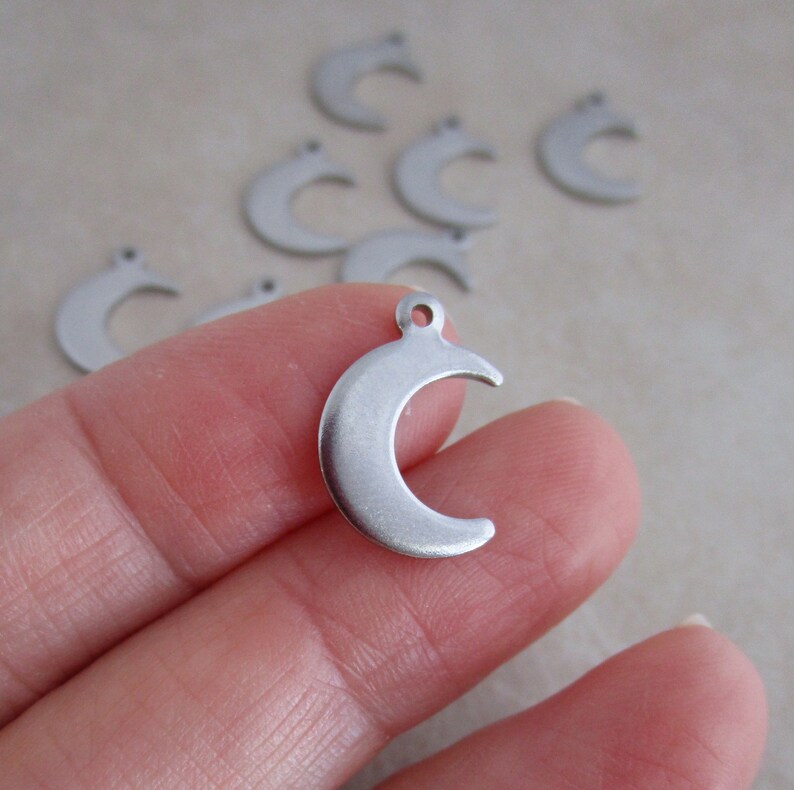 May include: A close-up of a silver crescent moon charm. The charm is held in a hand, and there are several other charms in the background.