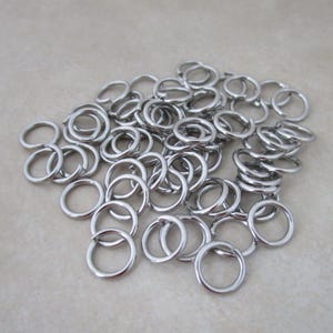 May include: A collection of numerous small, circular silver-toned metal jump rings. Each individual ring features a single cut or opening along its circumference, allowing for connection and assembly in various jewelry or craft projects.