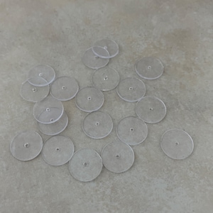 May include: A collection of clear, circular, flat plastic discs with a small hole in the center. The discs are scattered across a light gray surface. These small, transparent components are likely used for crafting or jewelry making.