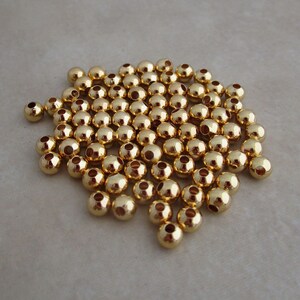 May include: A collection of small, shiny, gold-colored beads. Each bead is round with a hole running through the center. The beads are scattered across a neutral-toned surface, reflecting light and creating a bright, metallic appearance.