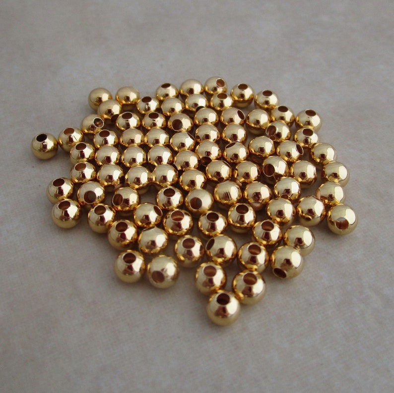 May include: A collection of small, shiny, gold-colored beads. Each bead is round with a hole running through the center. The beads are scattered across a neutral-toned surface, reflecting light and creating a bright, metallic appearance.