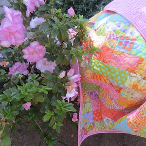 May include: A pink and green patchwork quilt with a pink floral print backing. The quilt is folded and lying on a bed of pink azalea flowers.