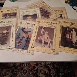 May include: A collection of eight Magic Threads craft pattern envelopes featuring illustrations of various dolls and figures. Each envelope is made of clear plastic and contains printed instructions and images of finished projects like gnomes, fairies, and dream catchers.