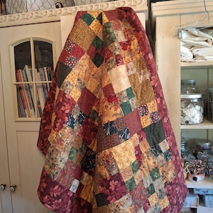 May include: A patchwork quilt featuring a variety of square fabric blocks in shades of burgundy, forest green, mustard yellow, and cream. The quilt displays diverse patterns including floral prints, leaf motifs, and textured solids stitched together.
