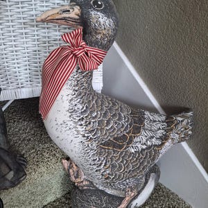May include: A decorative goose figurine with a detailed feather pattern in shades of gray, brown, and white. It features a red and white striped bow around its neck. The goose is perched on a green carpeted surface.