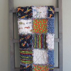 May include: A patchwork quilt with a variety of patterned squares, including airplanes, kiwi slices, and stripes. The quilt is displayed on a gray wooden ladder, showcasing its colorful design and textured edges.