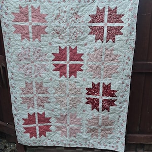 May include: A rectangular quilt featuring a repeating star pattern in various shades of red and pink. The quilt has a cream-colored background with a delicate floral print and a matching floral border. The stitching creates a textured surface.