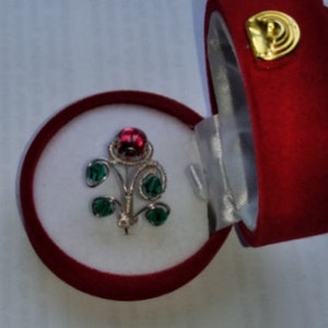 May include: A red and green flower brooch in a red velvet box. The flower is made of silver wire and has a red center and green leaves.