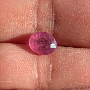 Peut inclure: Une pierre précieuse rose, de forme ovale et à facettes. La gemme a une surface polie avec de multiples facettes qui réfléchissent la lumière. La couleur est un rose vif, avec quelques inclusions internes visibles. La pierre est posée sur un fond de couleur chair.