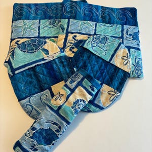May include: A quilted tote bag and matching oven mitts feature a sea life design. The bag and mitts are in shades of blue and tan, with images of turtles, crabs, and seashells. The bag has a wave pattern along the top edge.