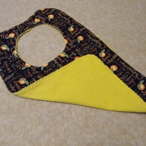 May include: A black and yellow fabric bib with a citrus theme. The bib has a yellow terry cloth backing and a black fabric front with a repeating pattern of oranges, lemons, and the words "Sweet", "Tangy", "Citrus", "Marmalade", "Orange Juice", and "Peel".