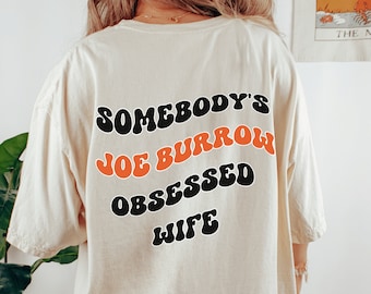 May include: A white t-shirt with the text "Somebody's Joe Burrow Obsessed Wife" printed on the back in black and orange letters.