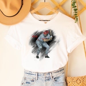 Op de afbeelding: Wit T-shirt met een aquarel illustratie van een honkbalspeler in slagpositie. De speler draagt een rode pet en blauw uniform. Het shirt wordt gecombineerd met een spijkerbroekshort, een strohoed en een geweven crossbodytas.