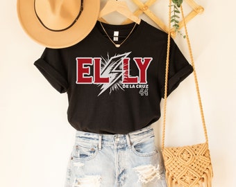 Può includere: Maglietta nera con una grafica rossa e bianca che dice "ELLY DE LA CRUZ 44". La maglietta è appesa a un appendiabiti di legno ed è abbinata a un cappello marrone e una borsa intrecciata.