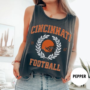 May include: A dark grey tank top with a white graphic of a football helmet with a wreath and the text "Cincinnati Football" with the numbers 19 and 67.