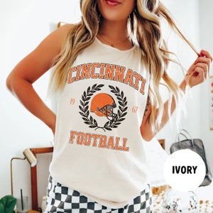 May include: A white tank top with a vintage-style graphic of a football helmet and the text "Cincinnati 19 67 Football" in black and orange. The tank top is on a model wearing black and white checkered shorts. The colour of the tank top is ivory.