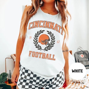 May include: White tank top with a vintage-style graphic of a football helmet and the text "Cincinnati Football" with the years 19 and 67 on either side of the graphic.