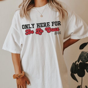 May include: White t-shirt with black text that reads "ONLY HERE FOR De La Cruz #44". The text "De La Cruz" is in red.