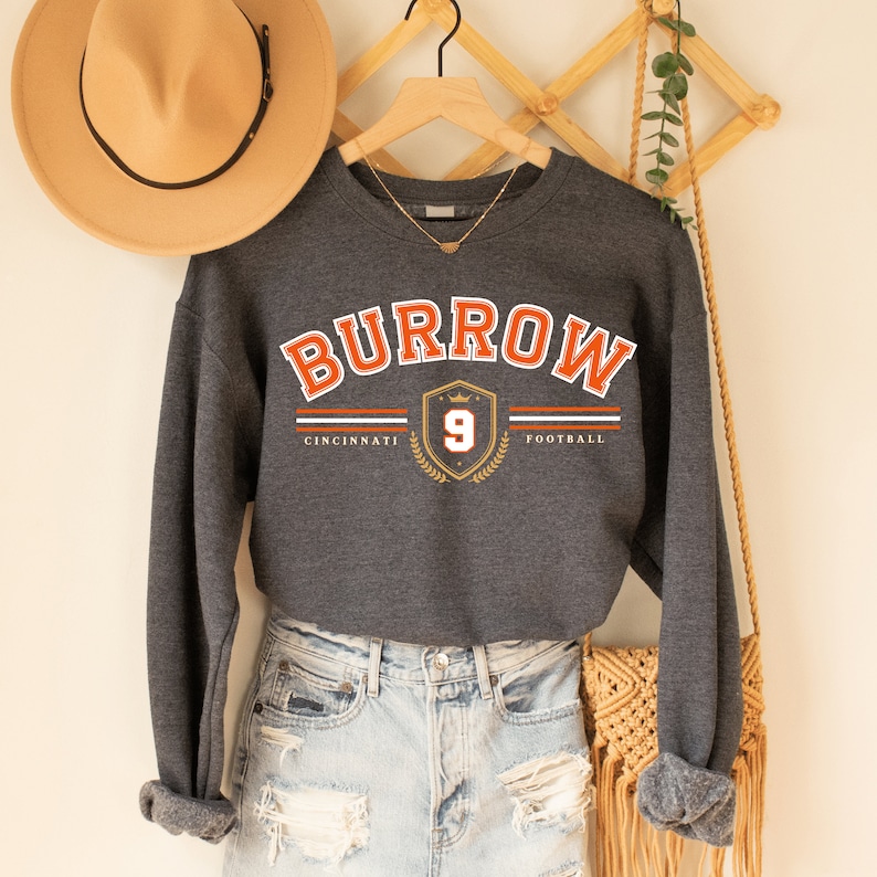 May include: A dark gray sweatshirt with the text "BURROW" in orange, a shield with the number "9" in gold, and the words "CINCINNATI" and "FOOTBALL" in white.