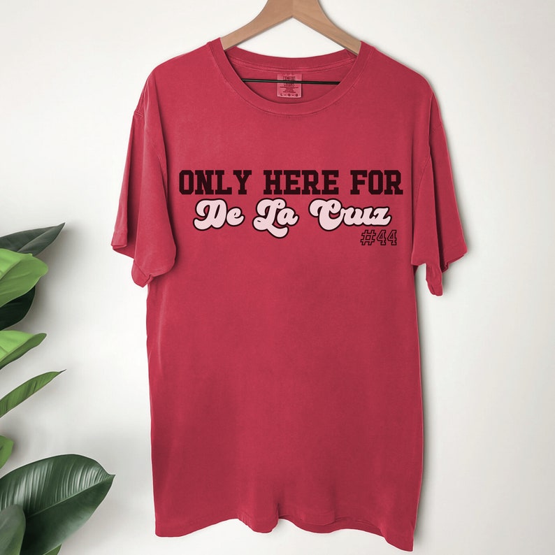 May include: Red t-shirt with the text "ONLY HERE FOR De La Cruz #44" in black and pink lettering.