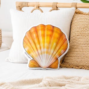 May include: A seashell-shaped decorative pillow with a vibrant orange and yellow gradient design. The pillow has a white border and the text "shell camp" is visible. The pillow is placed on a white bed with other pillows.