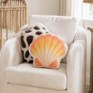 May include: A nursery with a light color palette features a rattan crib, a white armchair with shell and cow print pillows, and a framed beach scene. A woven lamp and a shelf with decor complete the coastal theme.