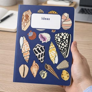 Może przedstawiać: Niebieski notatnik ze słowem "Ideas" w białym prostokącie. Okładka ozdobiona jest różnymi ilustracjami muszli w odcieniach brązu, bieli, żółci i czerni. Notatnik trzymany jest przez osobę.