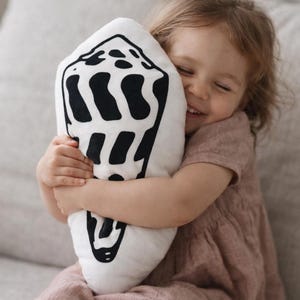 May include: A white pillow with a black seashell design is held by a young child. The pillow is shaped like a seashell and has a simple, graphic pattern. The child is wearing a pink dress.