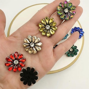 May include: Assortment of colorful flower-shaped embellishments with rhinestones. The embellishments are in various colors, including red, green, blue, yellow, and black.