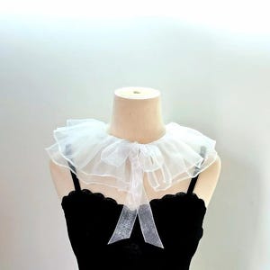 May include: A white sheer organza ruffled collar featuring a layered design with a delicate tie closure at the center front. The collar is displayed on a mannequin wearing a black sleeveless top with a scalloped neckline edge.