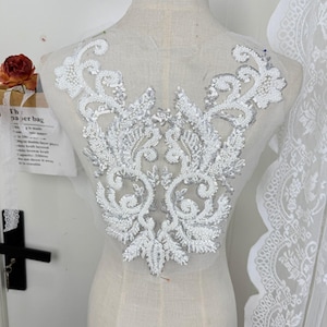 May include: A white and silver beaded applique with an ornate floral design. The applique is made of sequins and beads, and is attached to a sheer fabric backing. The design is symmetrical and features swirling patterns.